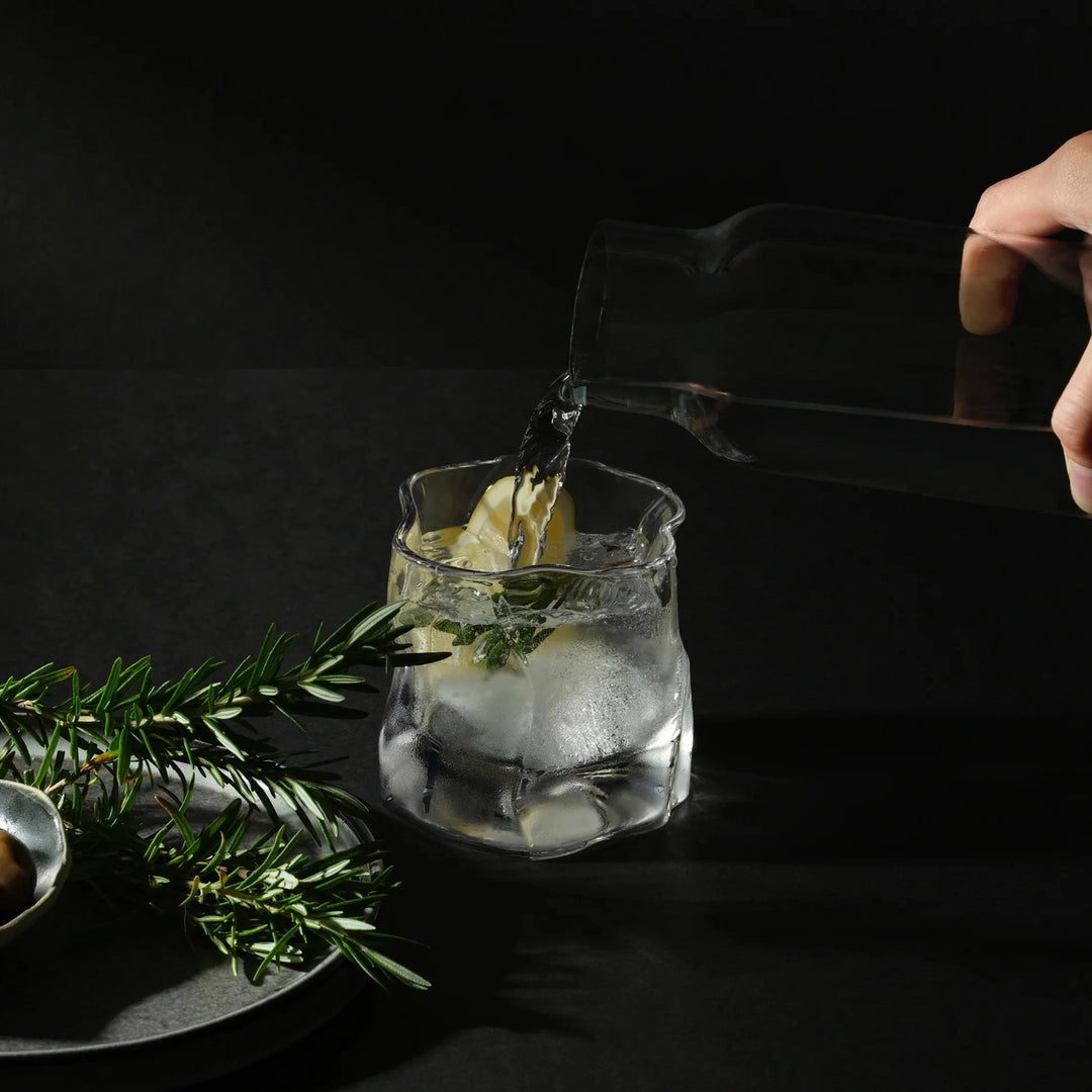 Person pouring a clear liquid into a glass with ice and herbs on a dark background
