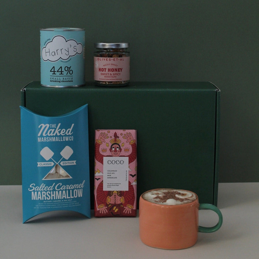 Hot chocolate in a mug in front of gift hamper with contents including hot chocolate in a tin, marshmallows, nuts and chocolate