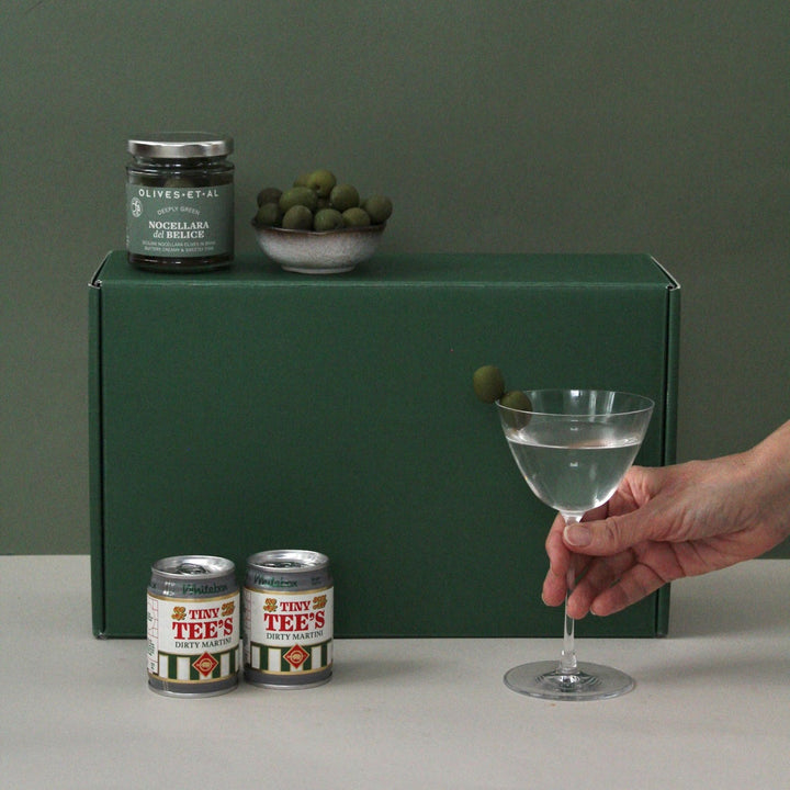 Person holding a cocktail with olives, cans of martini up against a green himper giftbox