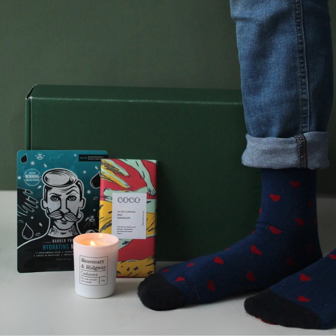 Person wearing dark blue socks with red hearts, standing next to a green box, colorful packaging, and a lit candle on a gray surface.